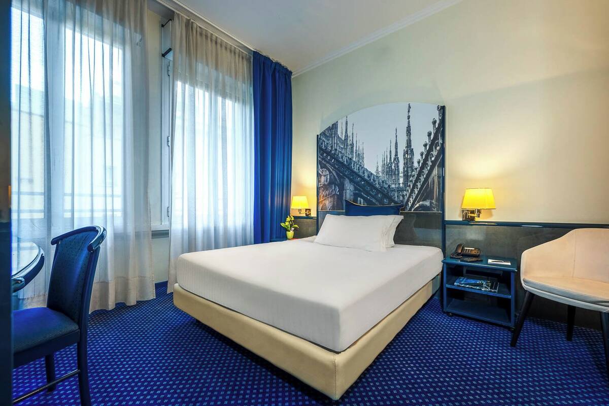 A hotel room with a large bed, a desk, a chair, and a painting of a cityscape on the wall.