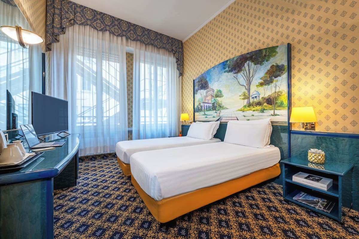 A hotel room with two beds, a desk with a laptop, a TV, a painting on the wall, and a patterned carpet.
