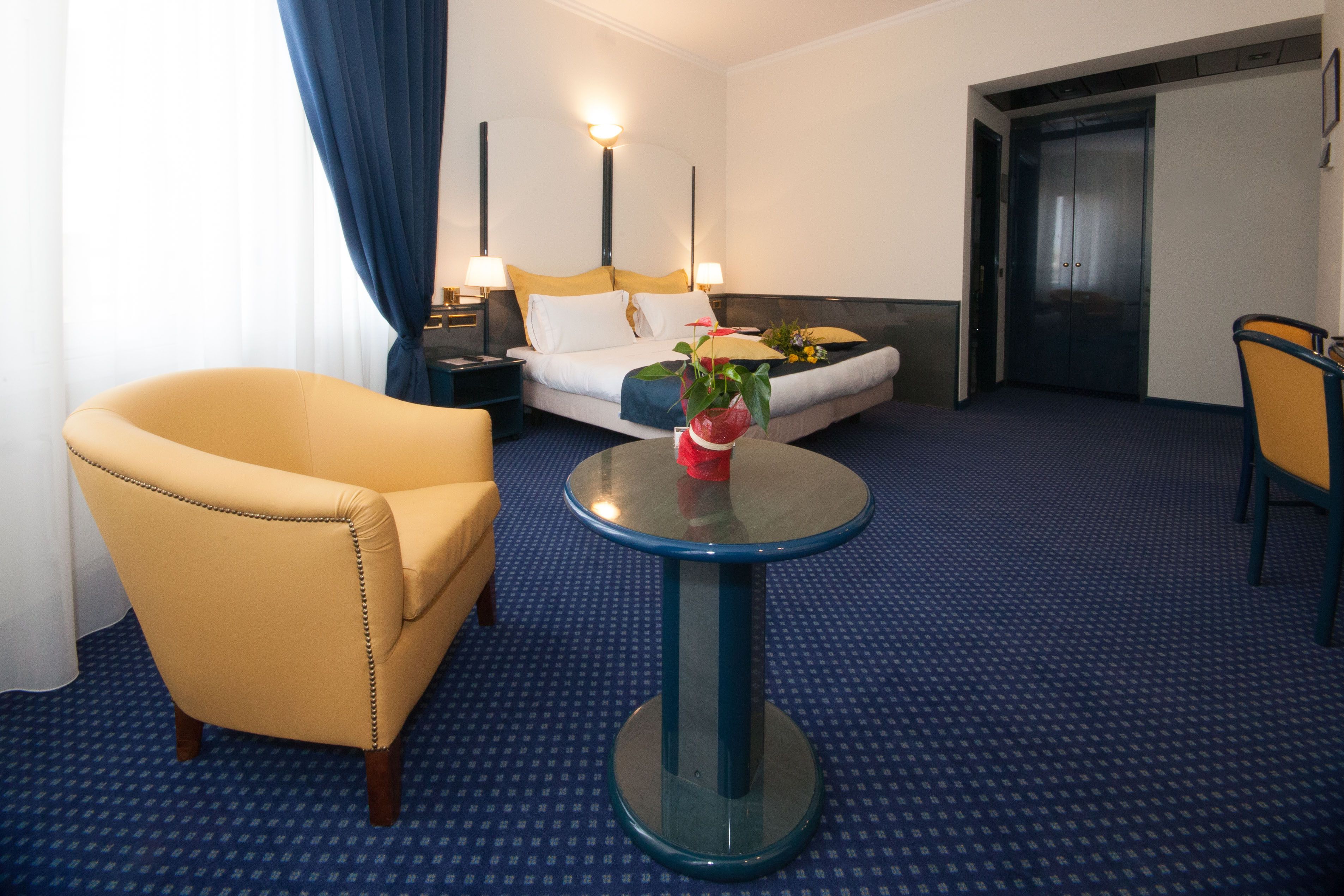 Habitació d'hotel amb un llit, una butaca groga, una taula rodona amb un ram i una catifes blava.