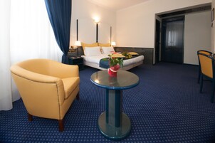 Een hotelkamer met een bed, een gele fauteuil, een ronde tafel met een vaas en een blauw tapijt.