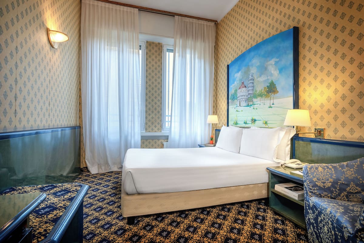 A hotel room with a large bed, a desk with a lamp, a chair, and a painting on the wall.