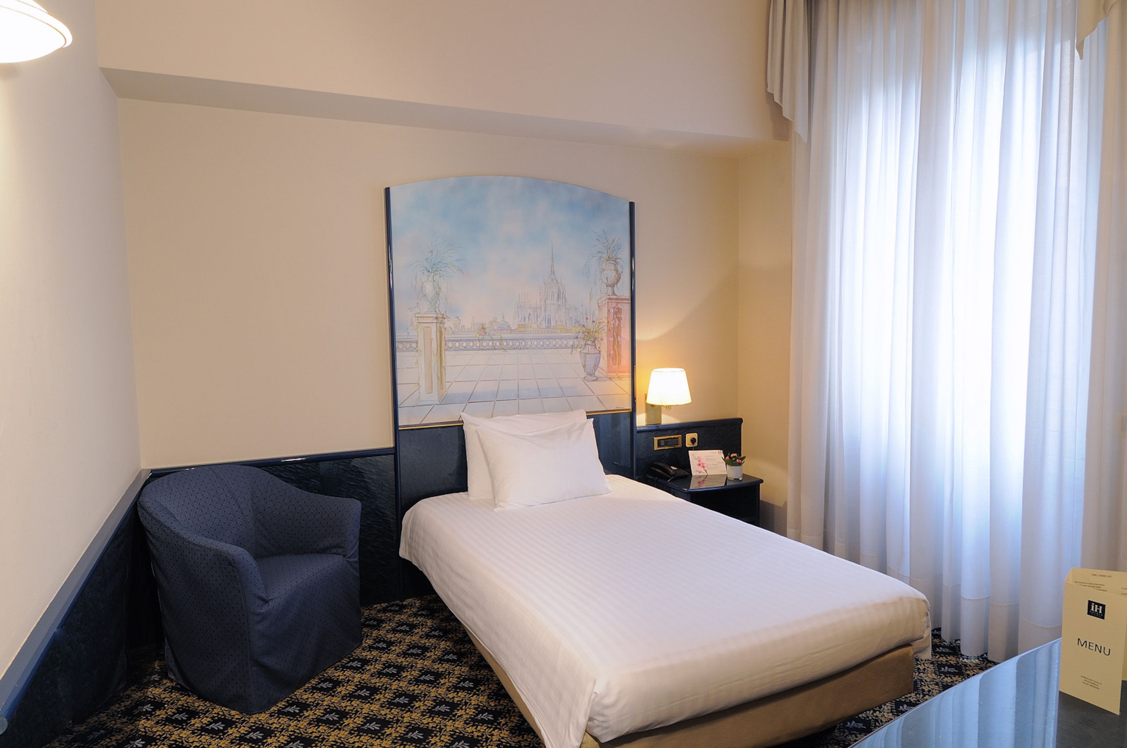 A hotel room with a bed, a chair, a nightstand with a lamp, a painting on the wall, and a window with curtains.