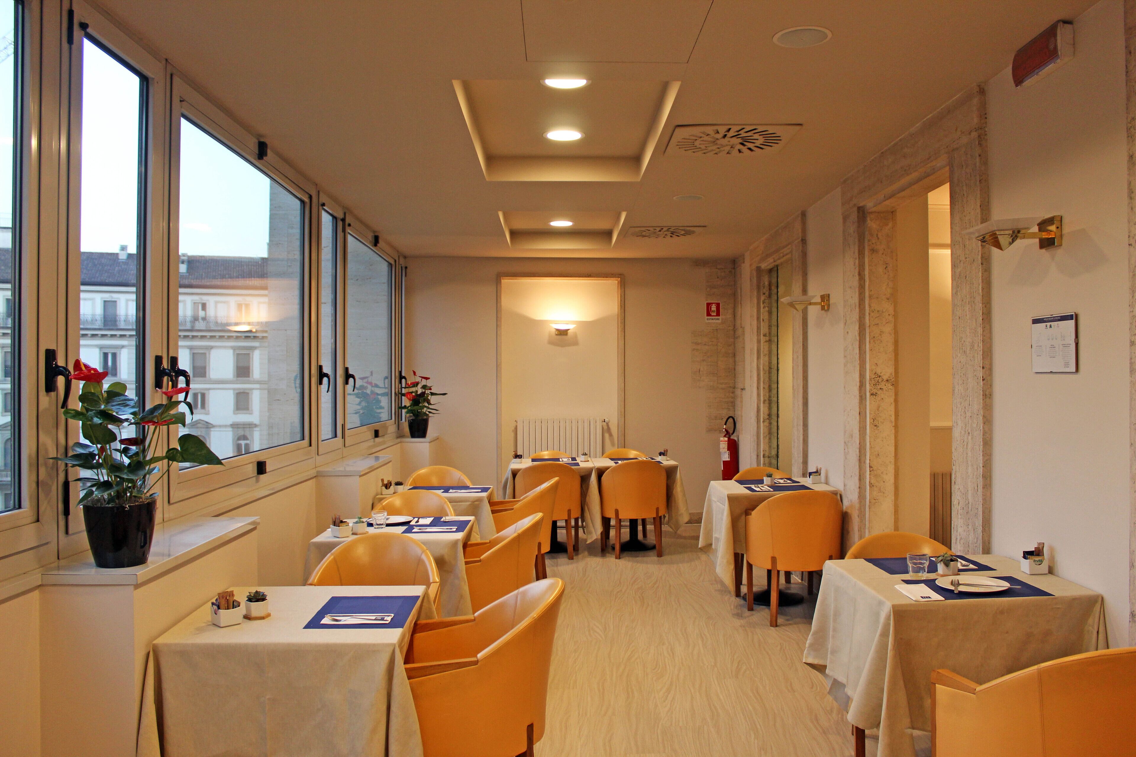 A hotel dining area with tables and chairs, large windows, and a view of buildings outside.