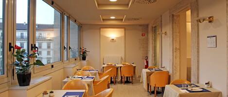 A hotel dining area with tables and chairs, large windows, and a view of buildings outside.