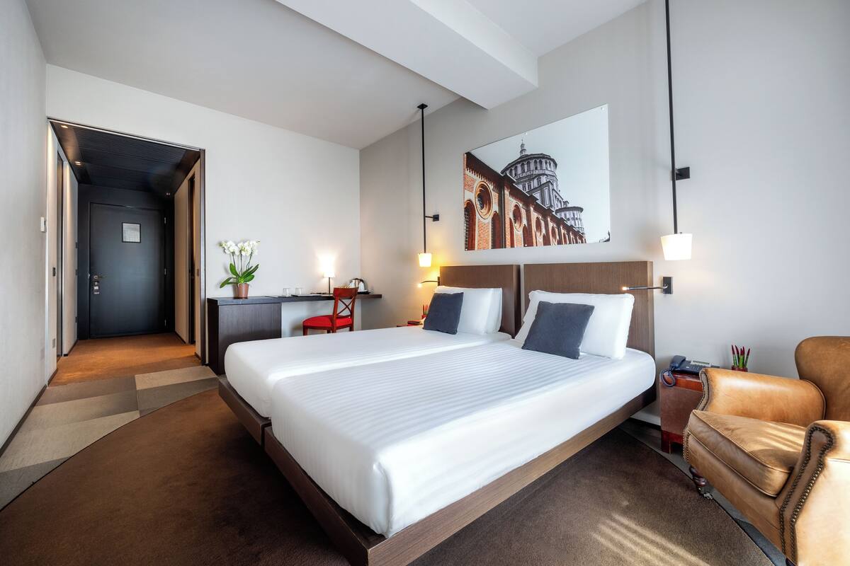 A hotel room with a large bed, a desk, a chair, a small table with a plant, and a wall-mounted lamp.