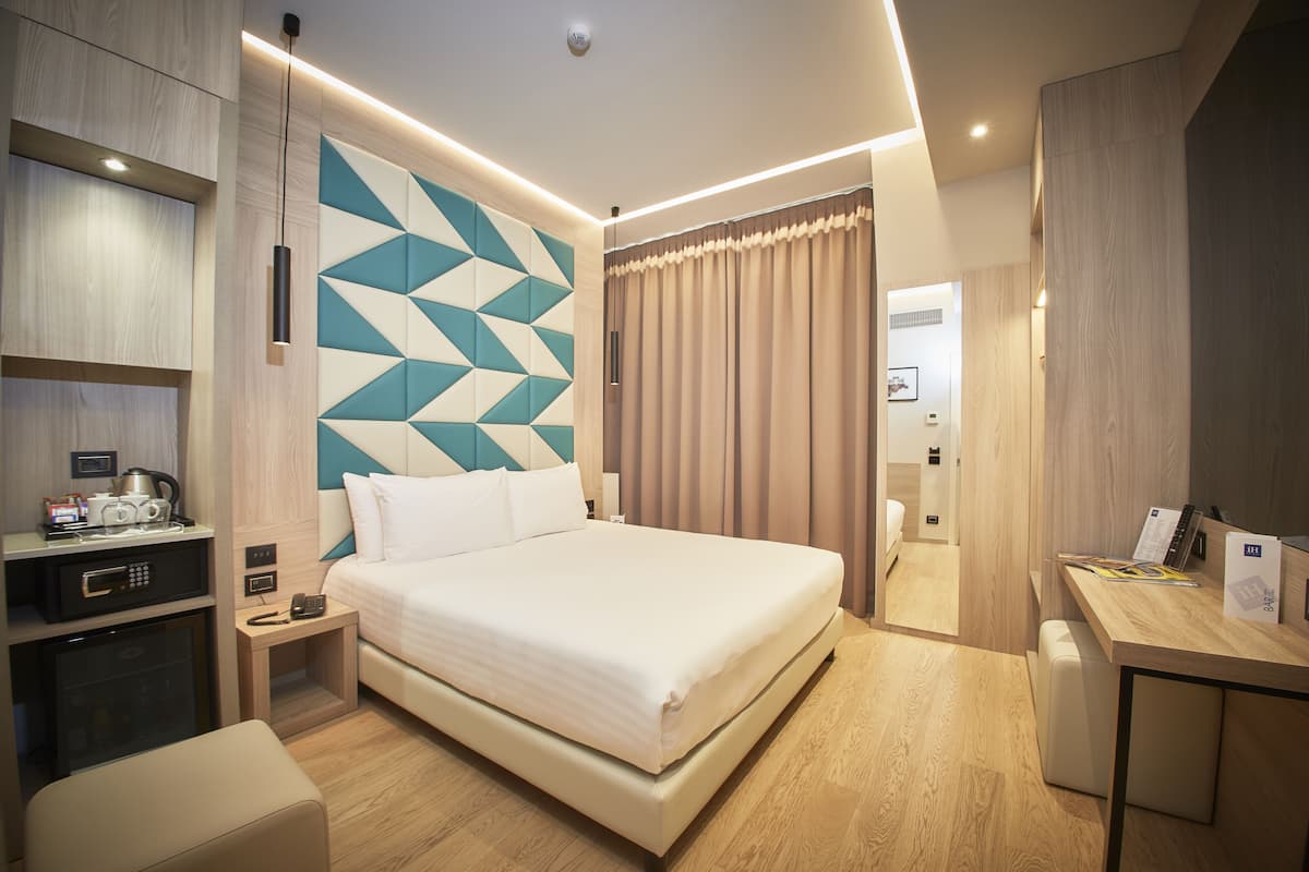 A modern hotel room with a large bed, bedside tables, a desk, and a kitchenette.