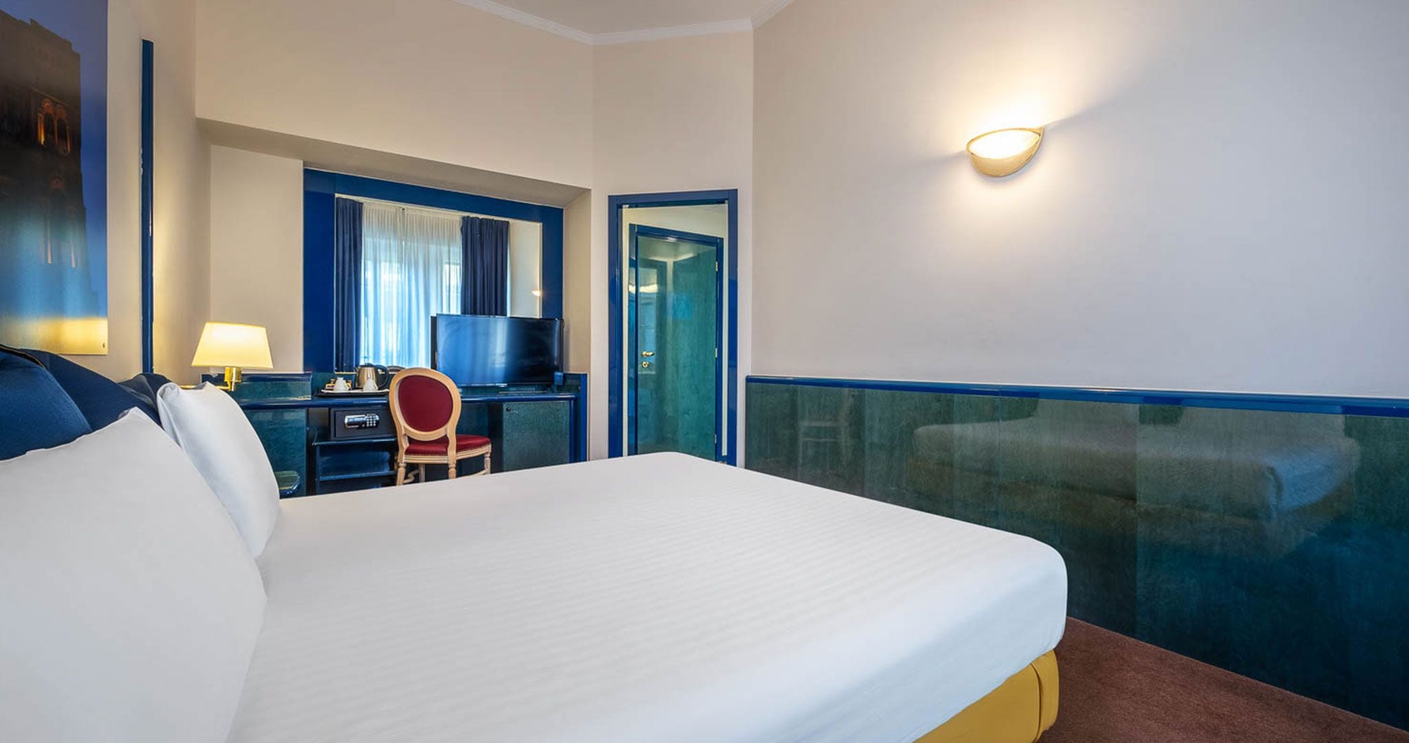 Een hotelkamer met een groot bed, een bureau met stoel, een televisie en een wandlamp.