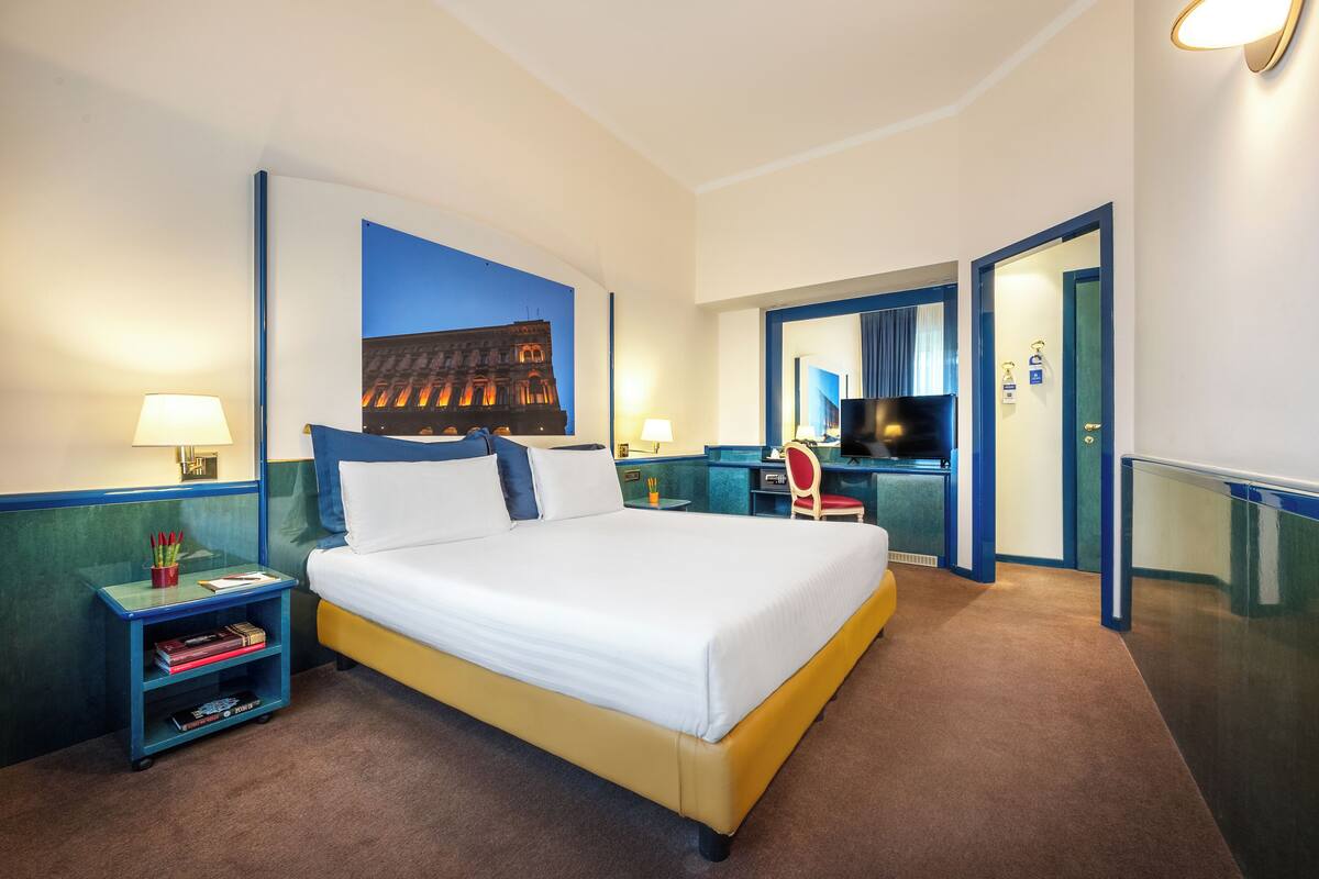 A hotel room with a bed, two bedside tables, a desk, a chair, a TV, and a bathroom door.