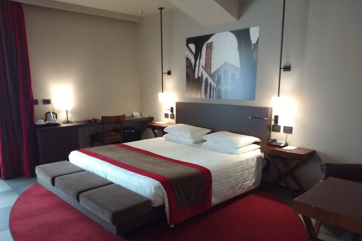 A hotel room with a large bed, a desk, a chair, and a red carpet.