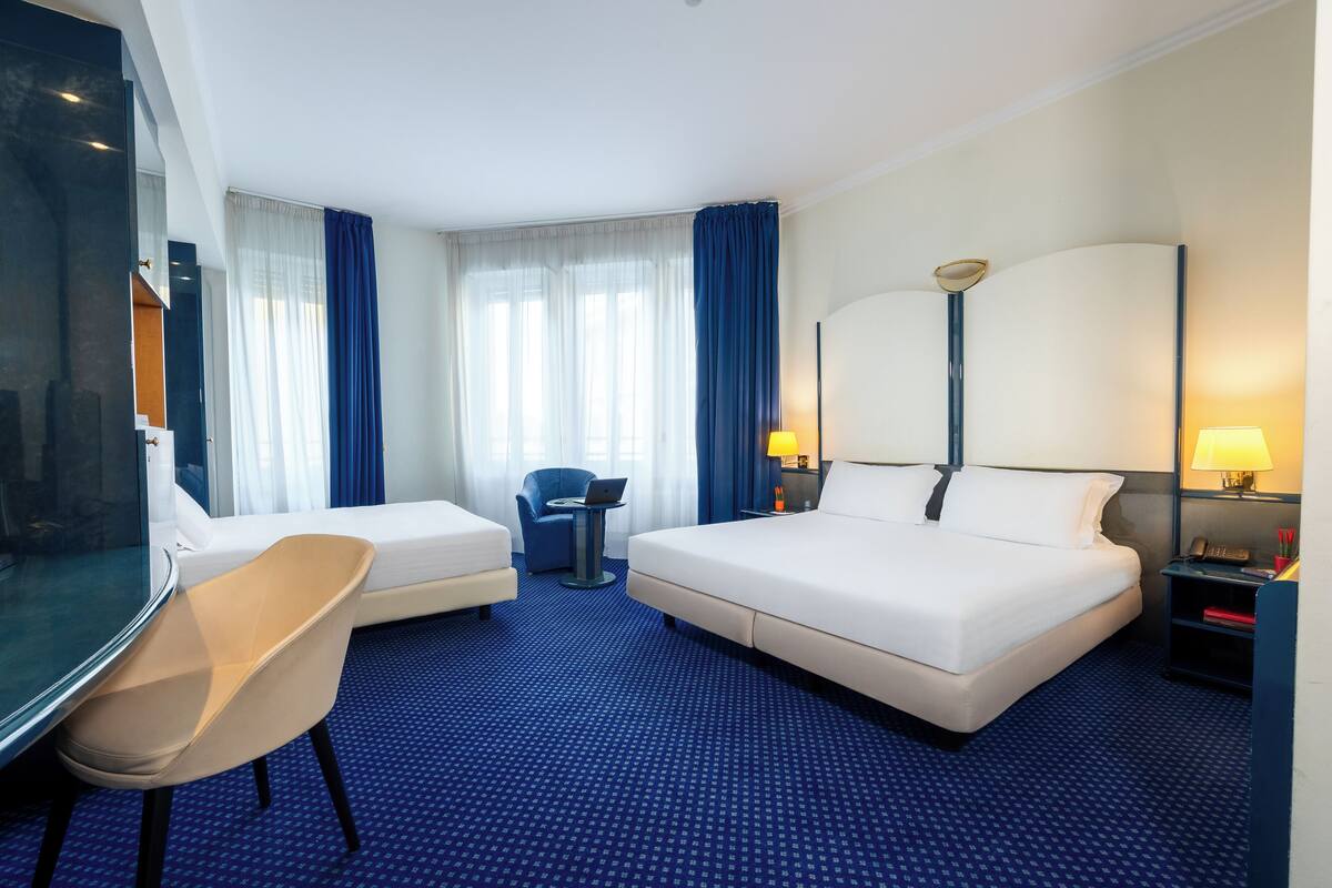 A hotel room with a large bed, a smaller bed, a desk, a chair, and a desk lamp.