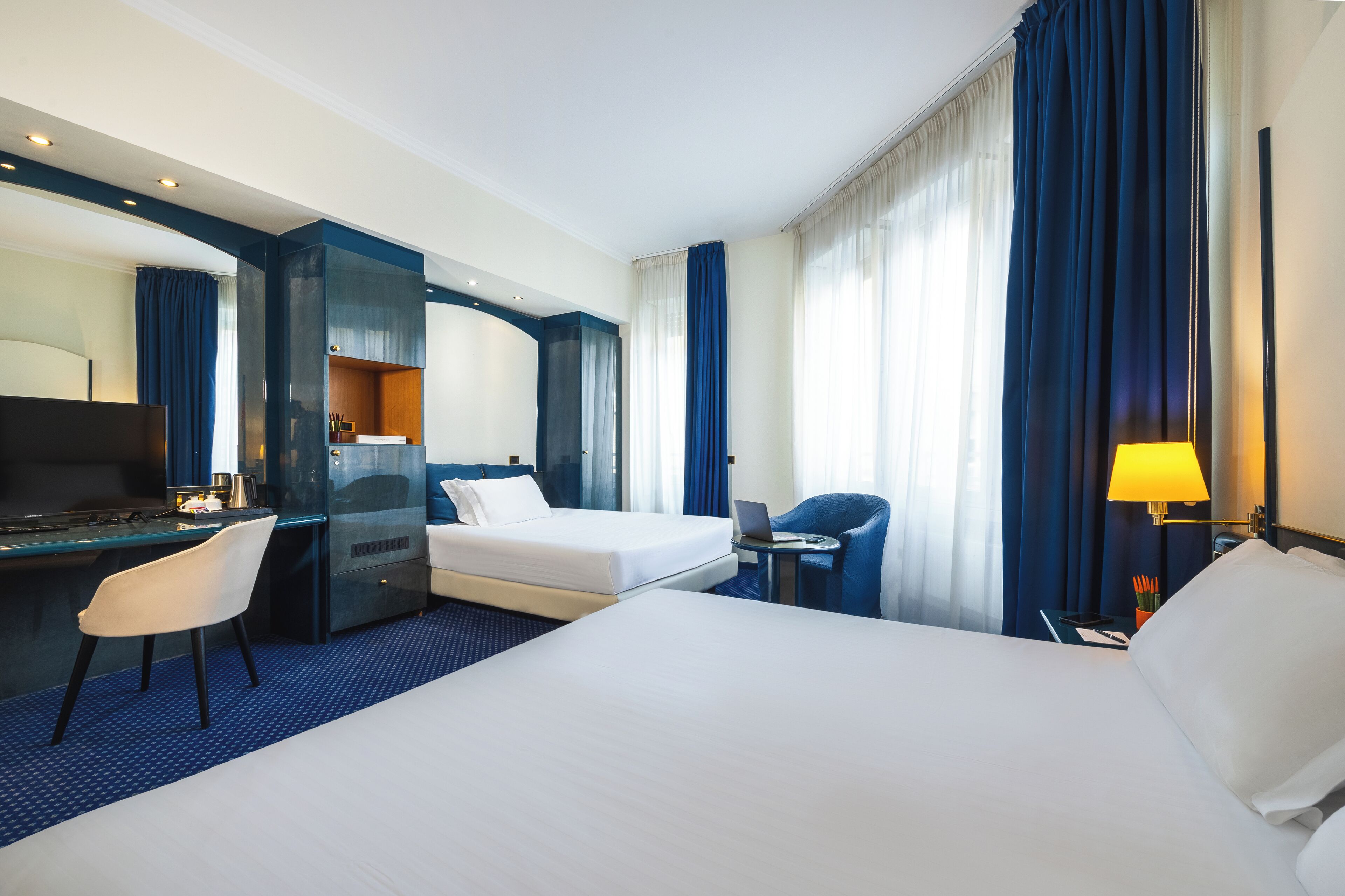 A hotel room with a large bed, a desk with a laptop, a chair, and a desk lamp.