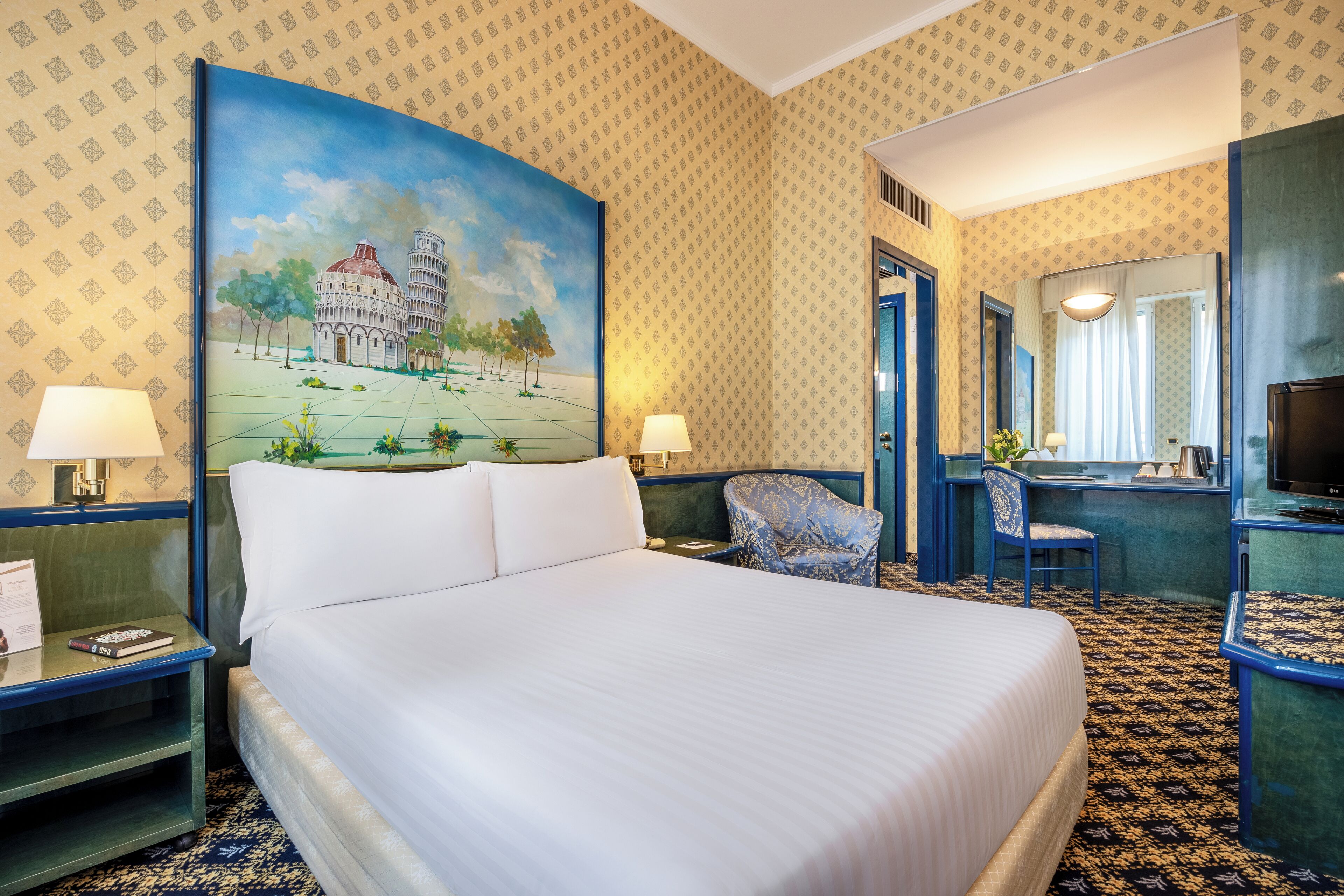 A hotel room with a large bed, two bedside tables, a desk with a chair, a television, and a large painting of a building.