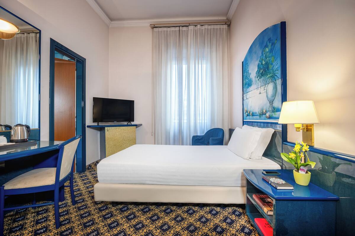 A hotel room with a bed, desk, chair, TV, and a painting on the wall.