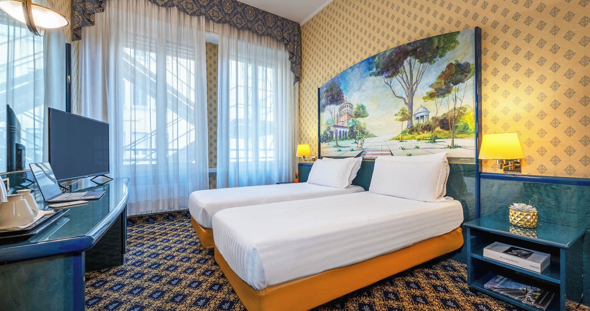 A hotel room with a bed, a desk with a laptop, a TV, and a painting on the wall.