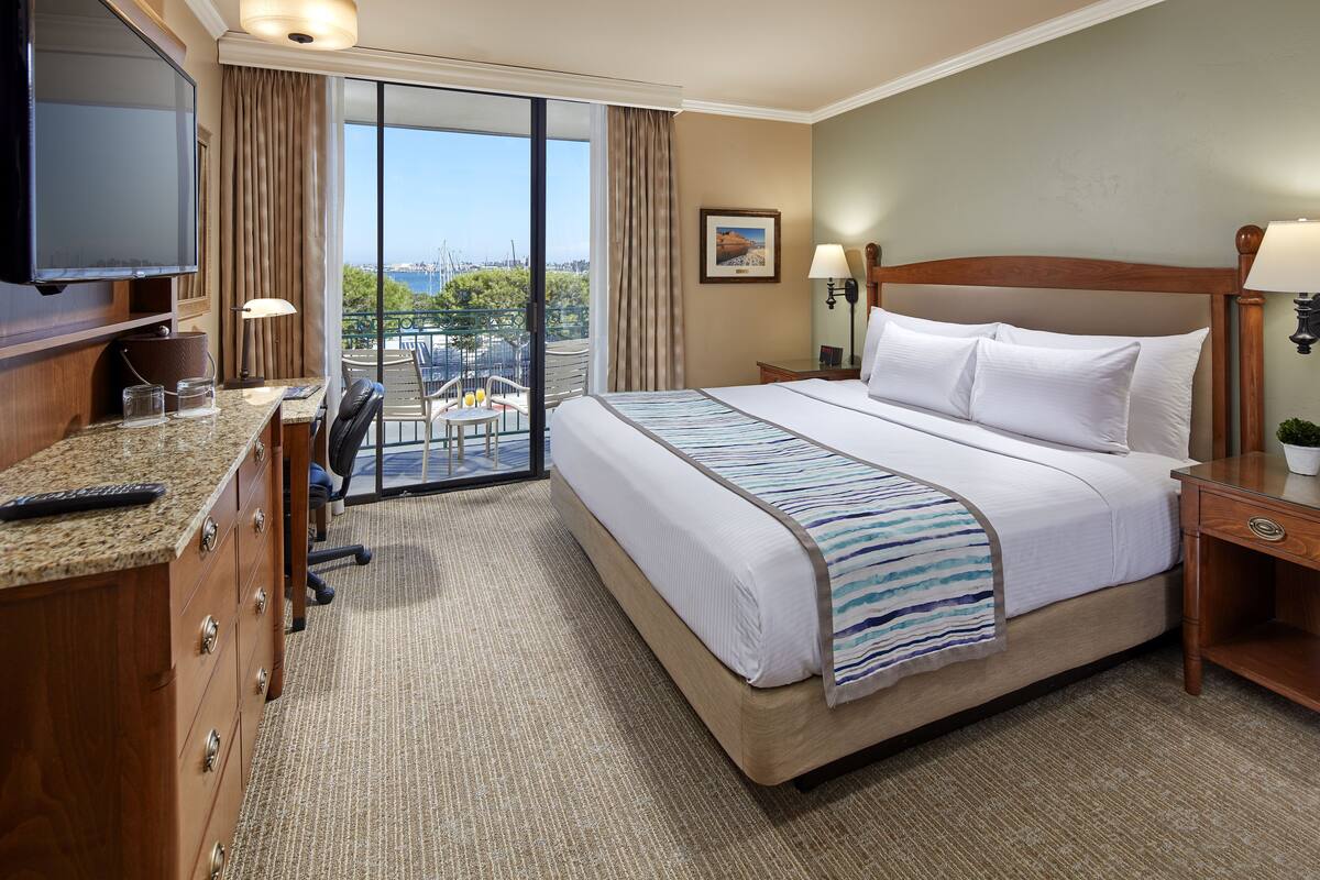 A hotel room with a large bed, a desk with a computer, a TV, and a balcony with a view of the city.