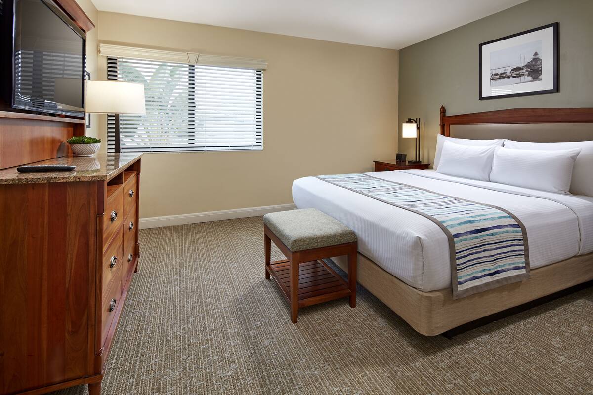 A hotel room with a bed, a TV, a desk with drawers, a lamp, and a window with blinds.