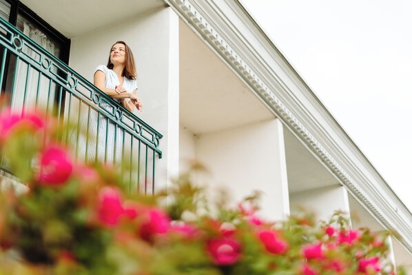 A person standing on a balcony with a railing, overlooking a building with a white facade and a decorative cornice.