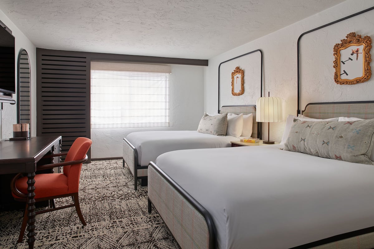 Une chambre d’hôtel avec deux lits, un bureau et une chaise. Il y a une fenêtre avec des stores, un miroir au mur et une horloge.