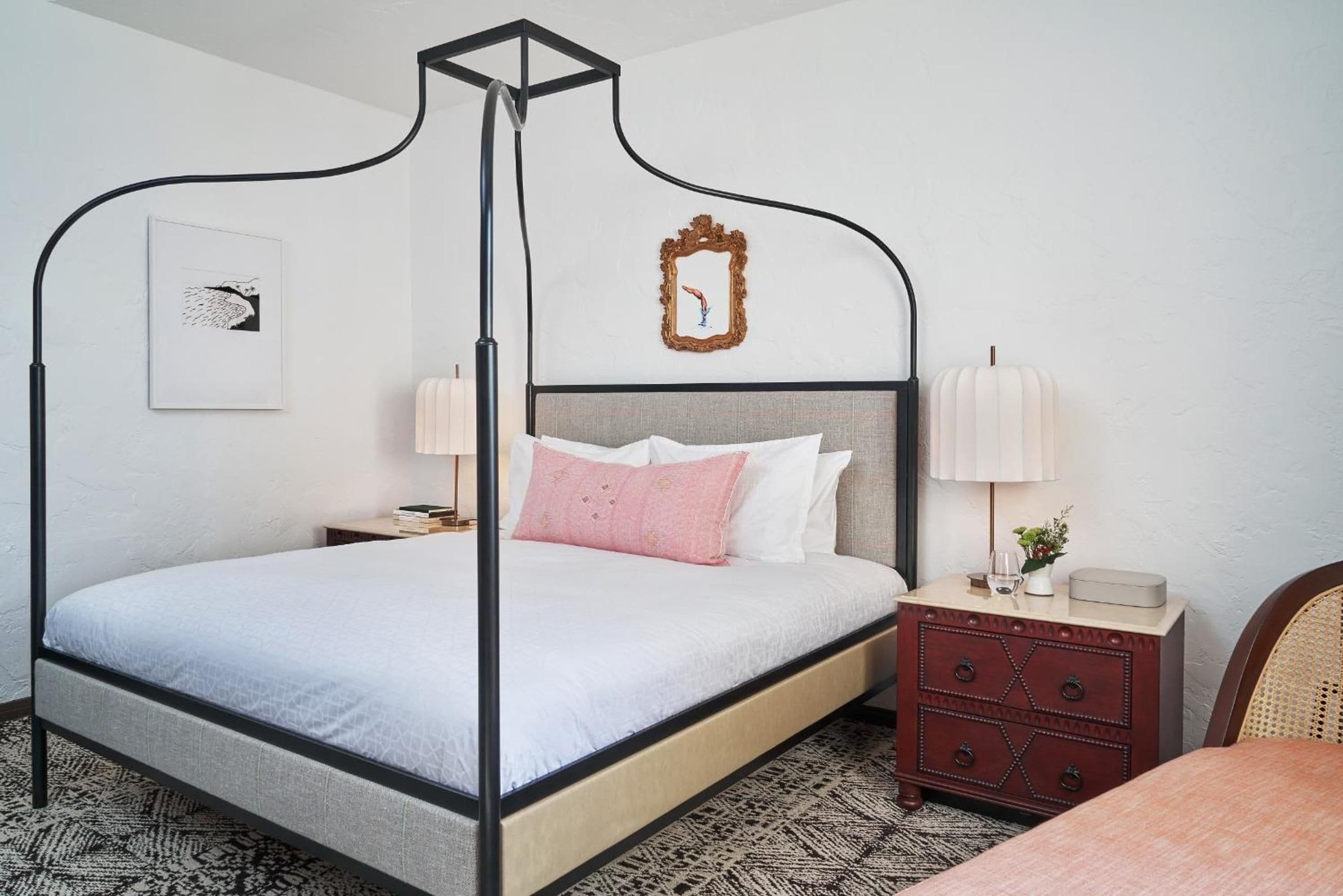 A four-poster bed with a headboard, a bedside table with a lamp, a small dresser, and a framed mirror on the wall.