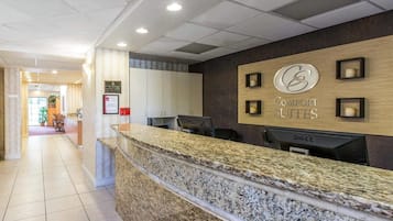 A hotel reception desk with a marble counter, a sign for 'Comfort Suites', and a view of the hallway leading to a dining area.