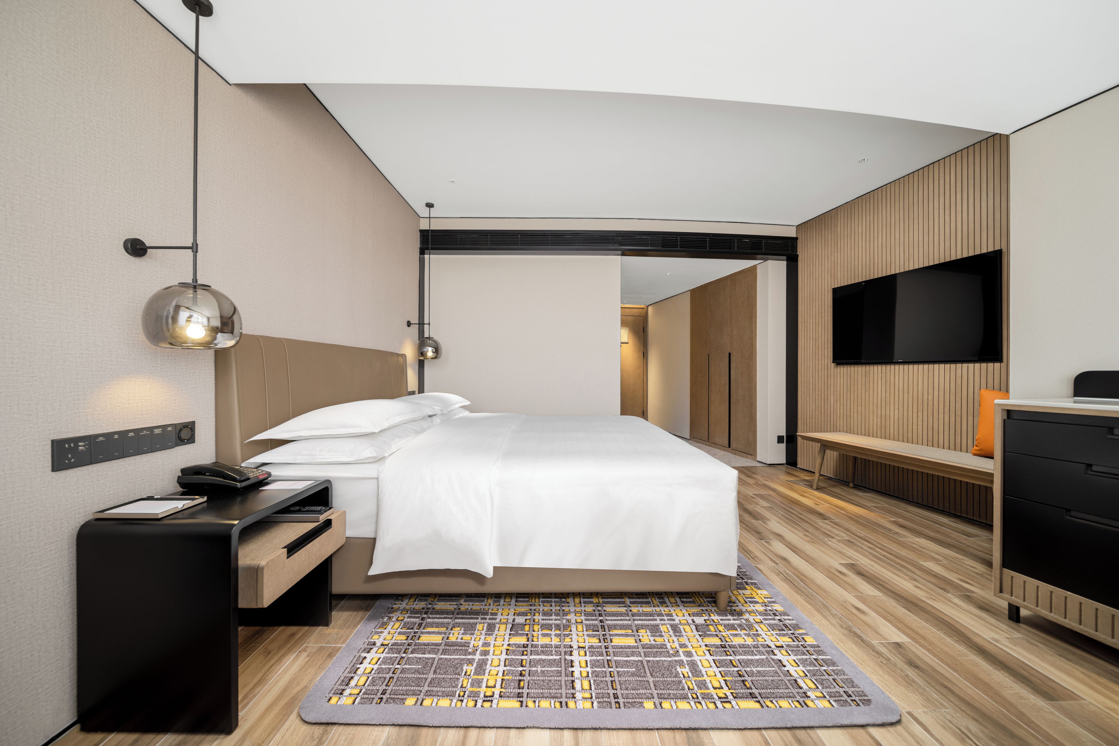 Une chambre d’hôtel moderne avec un lit bien fait, une table de chevet avec une lampe, un téléviseur à écran plat fixé au mur et un plancher en bois.