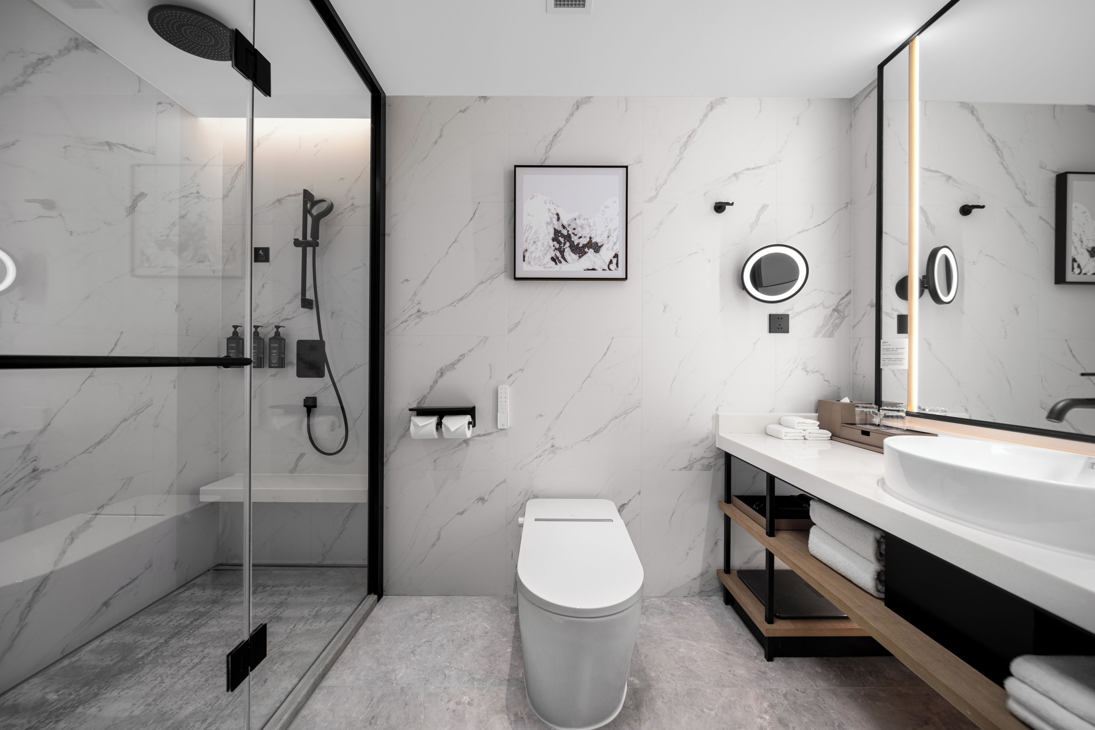Un baño moderno con ducha de vidrio, inodoro, lavamanos con un espejo grande y paredes de mármol.