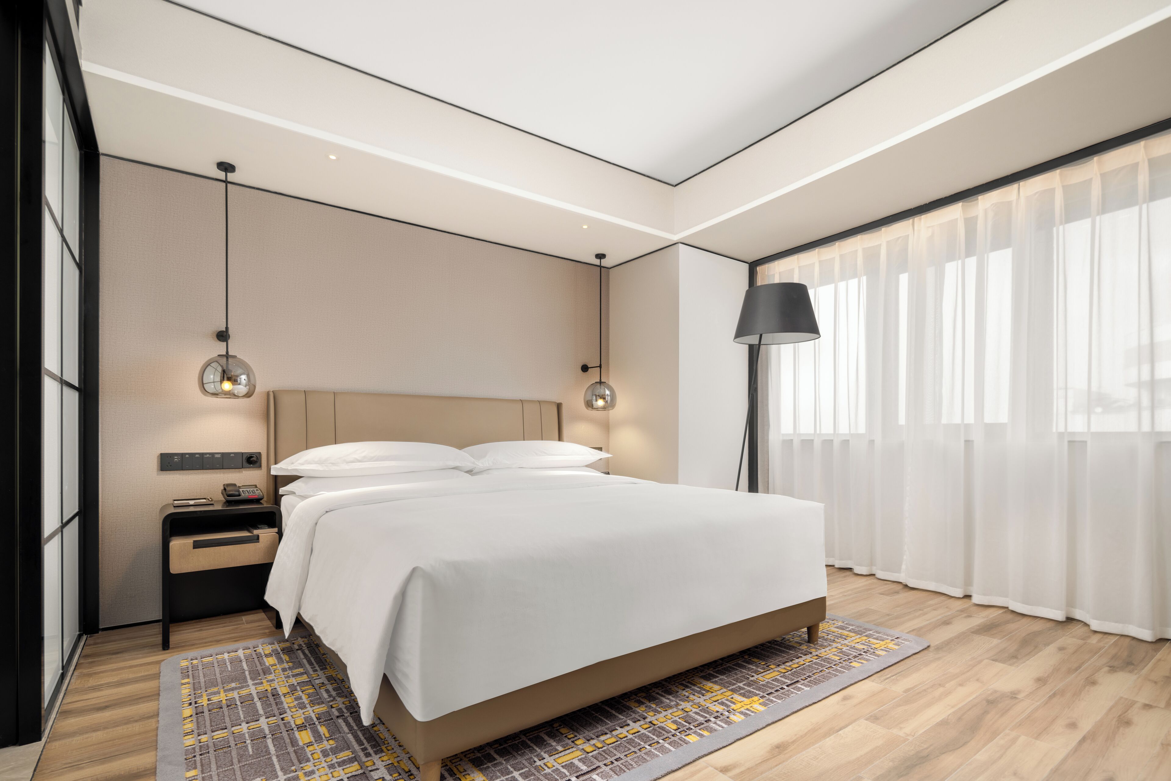 Een moderne hotelkamer met een groot bed, nachtkastjes, een lamp en uitzicht op het omliggende landschap door dunne gordijnen.