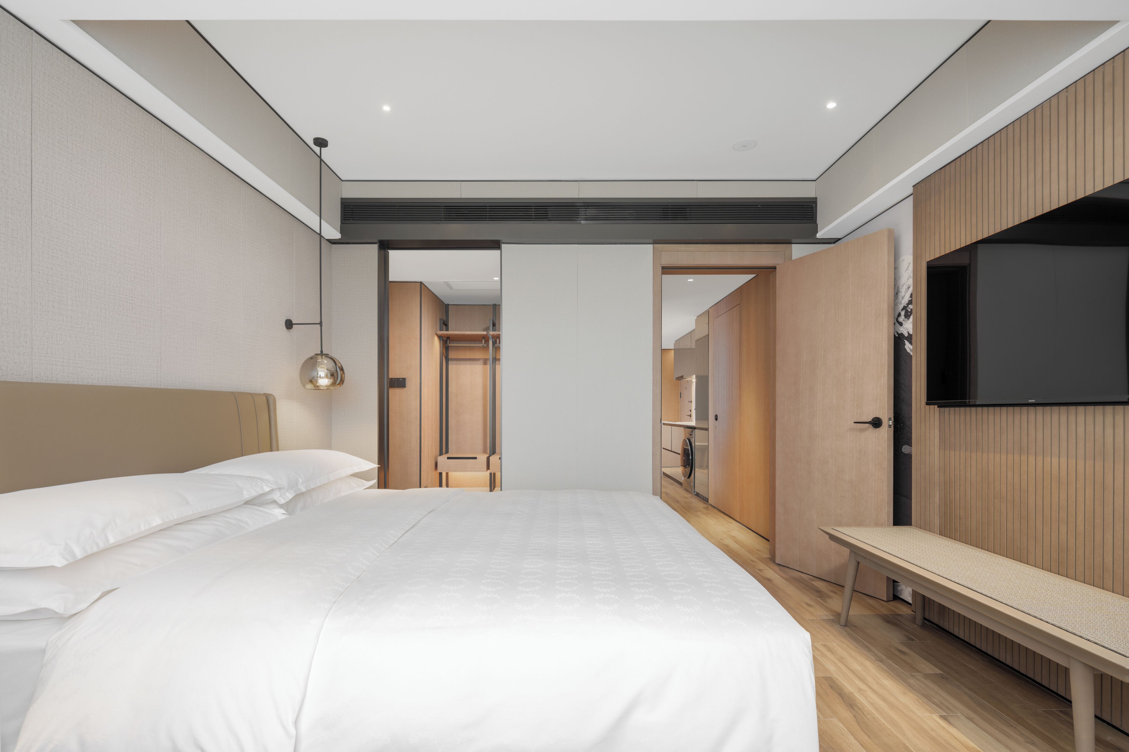 A modern hotel room with a large bed, a bench, a flat-screen TV mounted on the wall, and a walk-in closet.