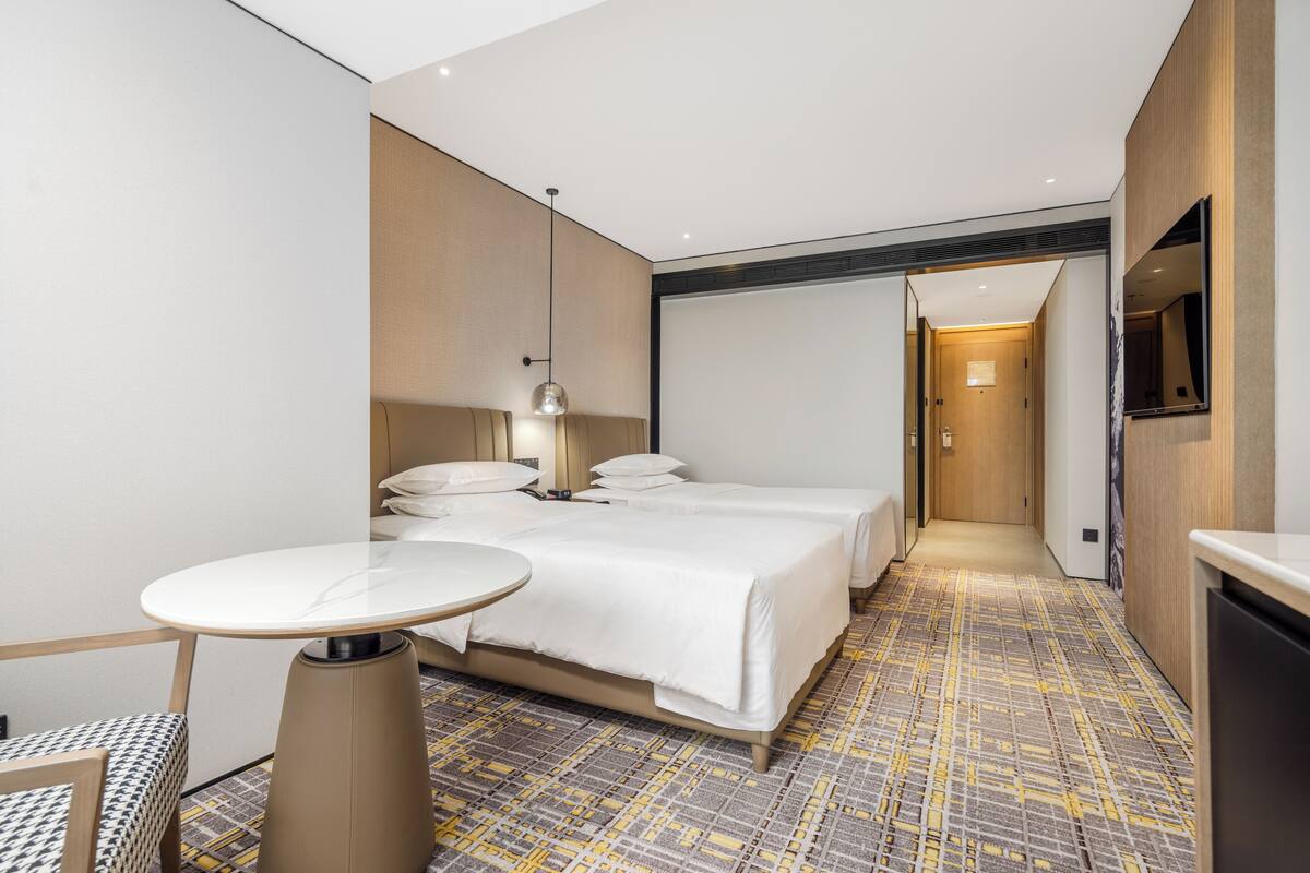 A modern hotel room with a large bed, a bedside table, a small round table, and a TV mounted on the wall.