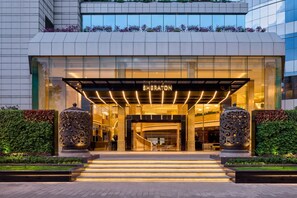 Ingresso di un hotel Sheraton dal design moderno, caratterizzato da una facciata in vetro, un ingresso ben illuminato e un'area verde con gradini che conducono alla porta.