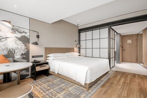 Una habitación de hotel moderna con una cama grande, un escritorio con silla, una mesita pequeña y un ventanal con vistas a la ciudad.