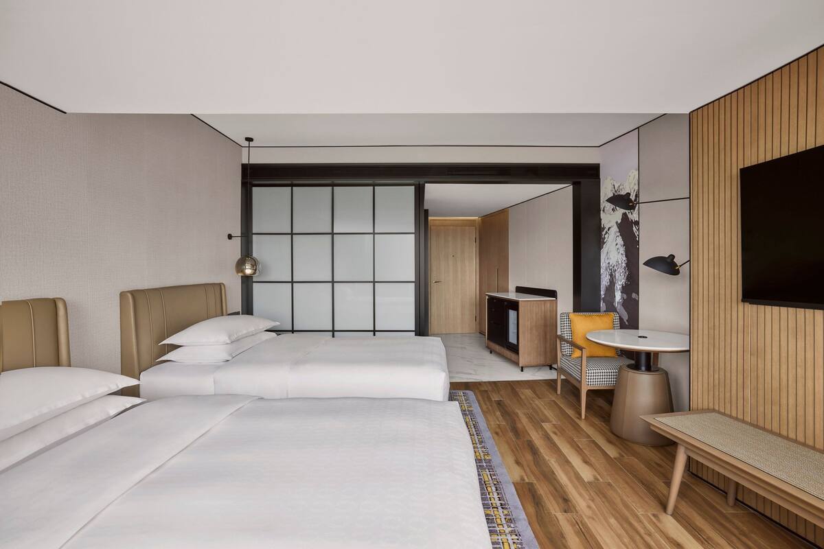 A modern hotel room with a large bed, a desk, a chair, a small table, and a TV.