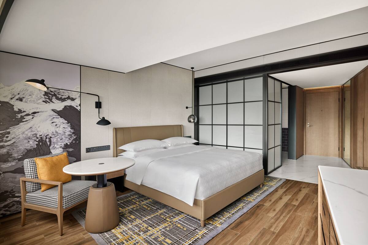 A modern hotel room with a large bed, a round table, a chair, and a wall-mounted lamp.