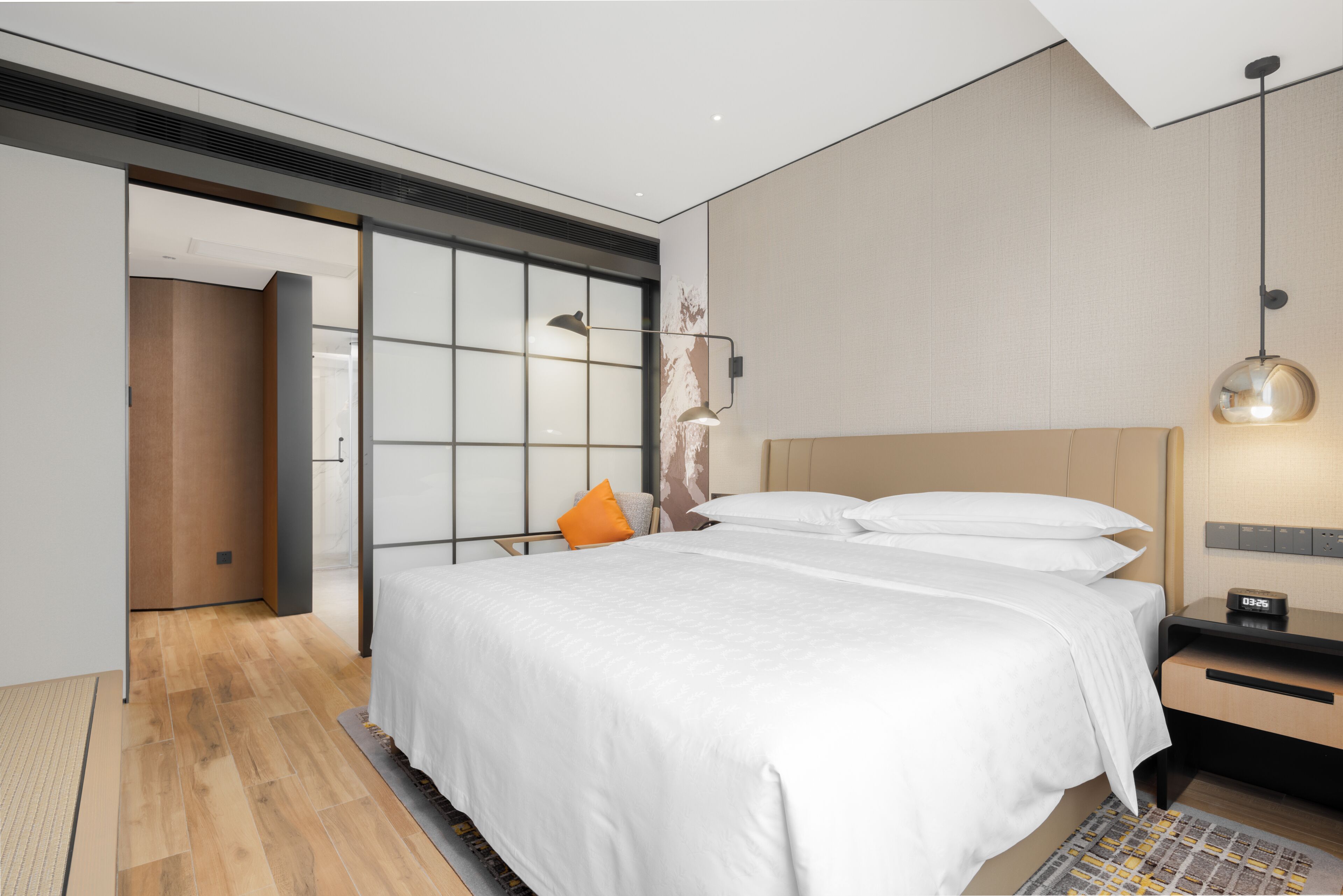 A modern hotel room with a large bed, a nightstand, a wall-mounted lamp, and a sliding door leading to another room.