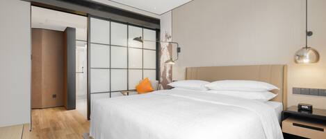 A modern hotel room with a large bed, a nightstand, a wall-mounted lamp, and a sliding door leading to another room.