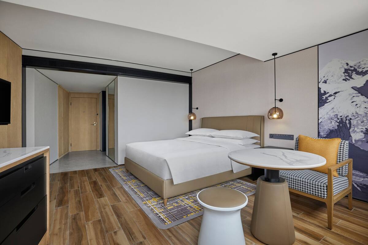 A modern hotel room with a bed, a round table, a chair, and a painting on the wall.