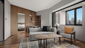 Une chambre d’hôtel moderne avec un canapé, un fauteuil et une petite table. On aperçoit un lit au fond et une fenêtre donnant sur la ville.
