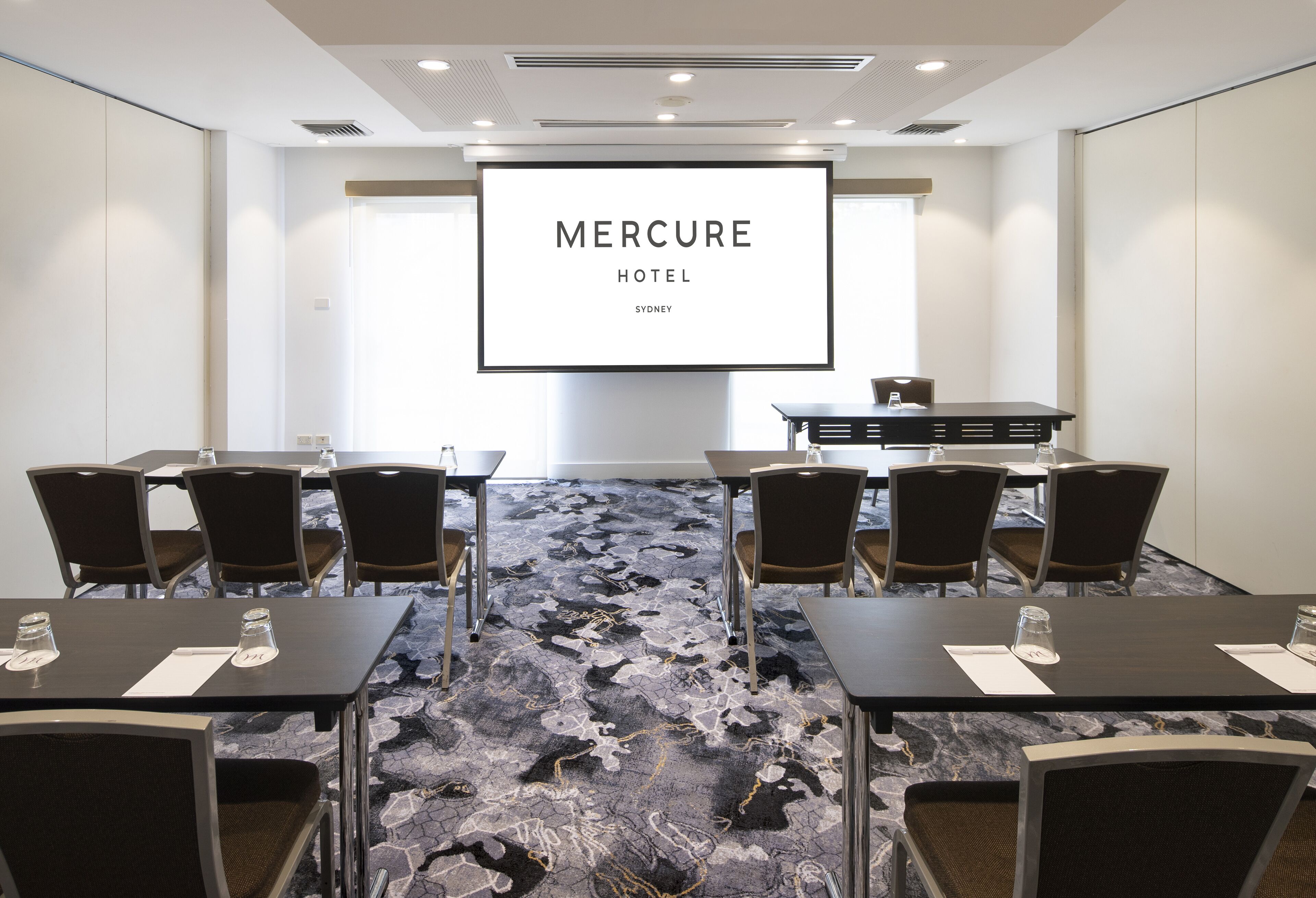 A conference room with a large screen displaying 'Mercure Hotel Sydney' and a carpeted floor.