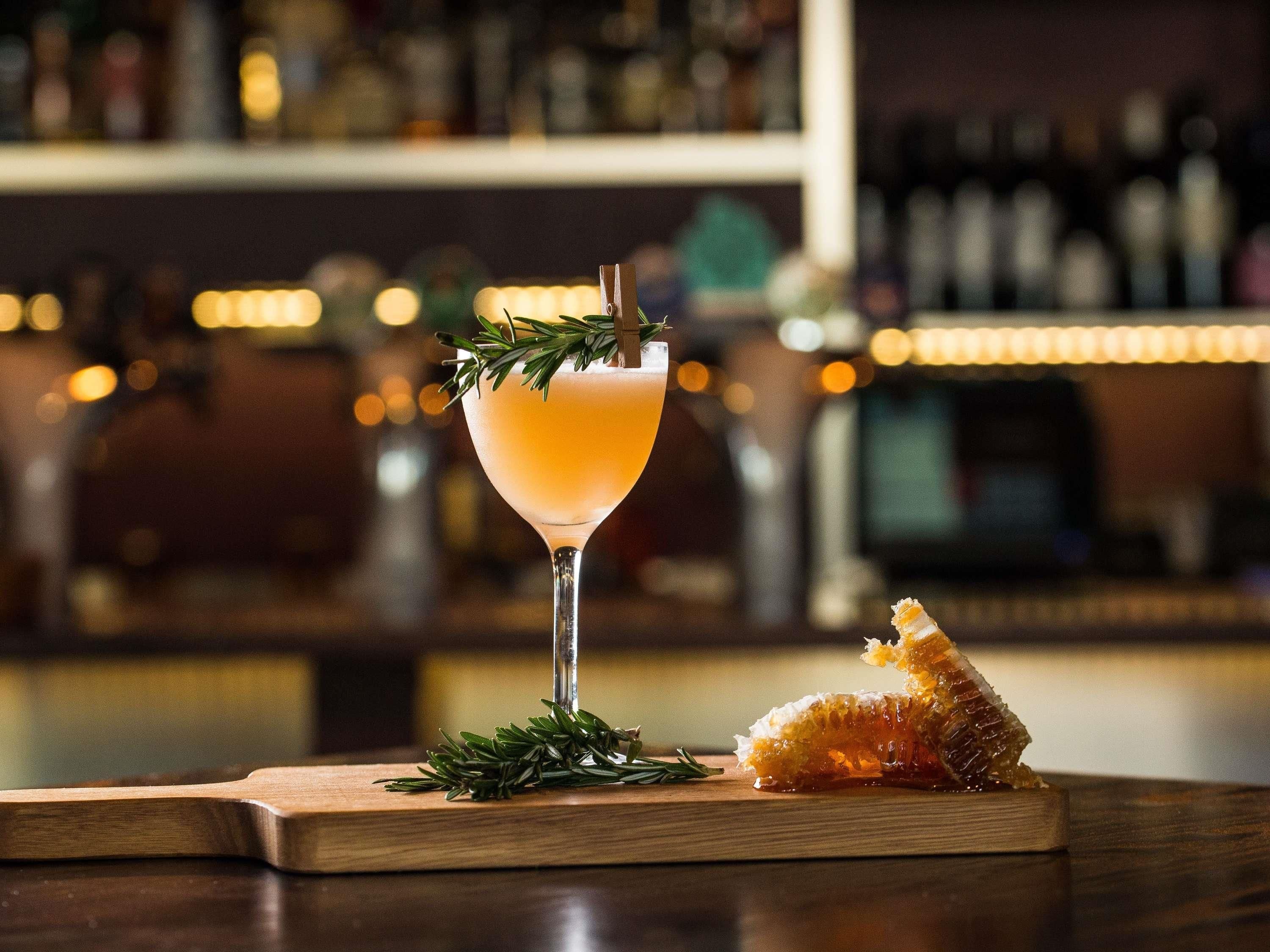 A cocktail glass with a garnish on a wooden bar counter, with a piece of honeycomb and rosemary nearby.