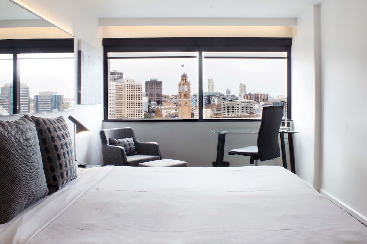 Une chambre d’hôtel avec une grande fenêtre, un lit, deux chaises et une petite table.