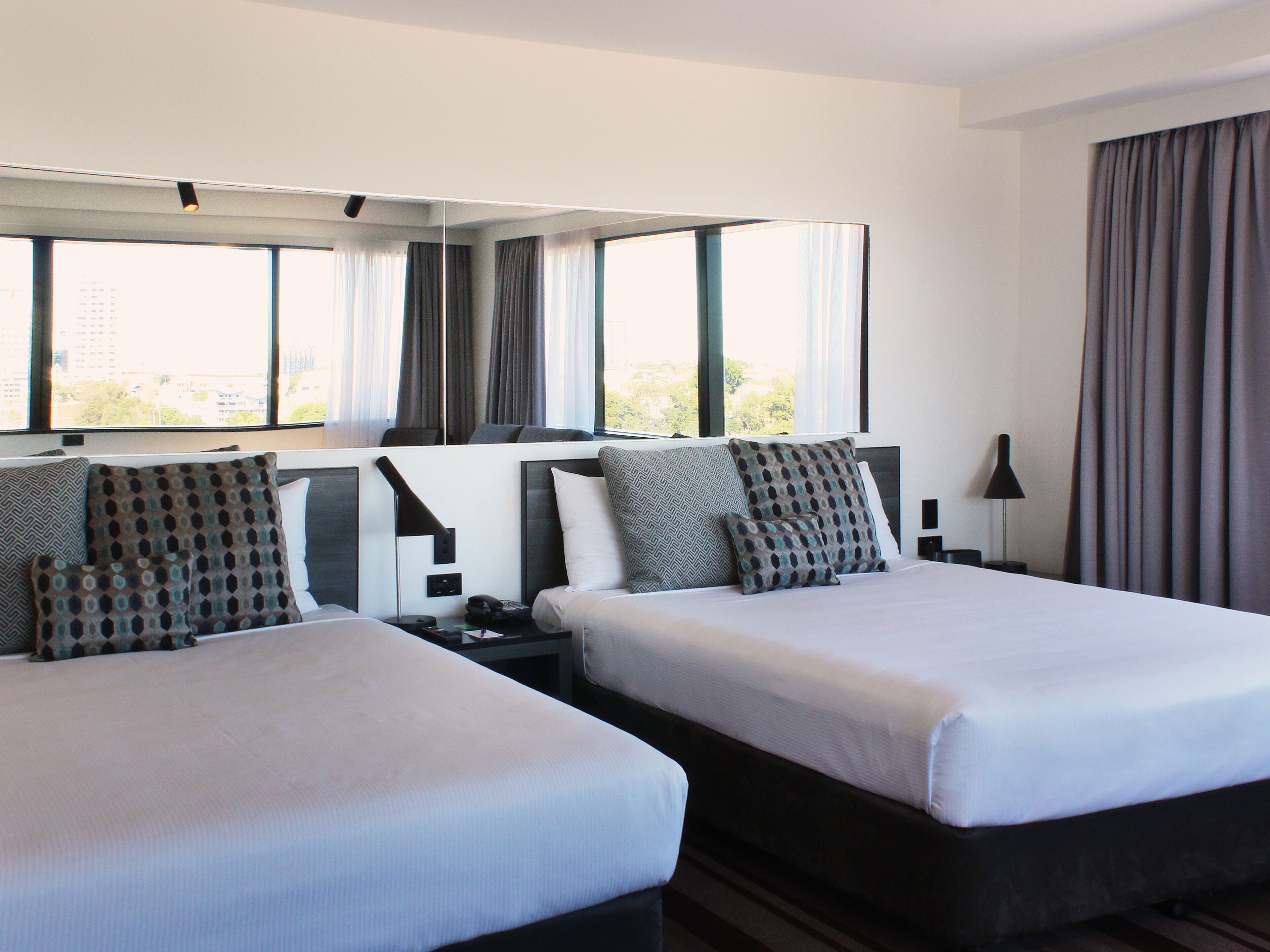 Ein Hotelzimmer mit zwei Betten, einem großen Fenster mit Vorhängen und Blick auf die Stadt.
