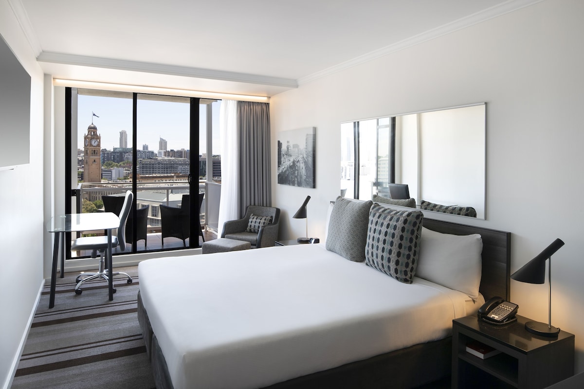 Une chambre d’hôtel avec un grand lit, un bureau et une chaise. La fenêtre offre une vue sur le paysage urbain.