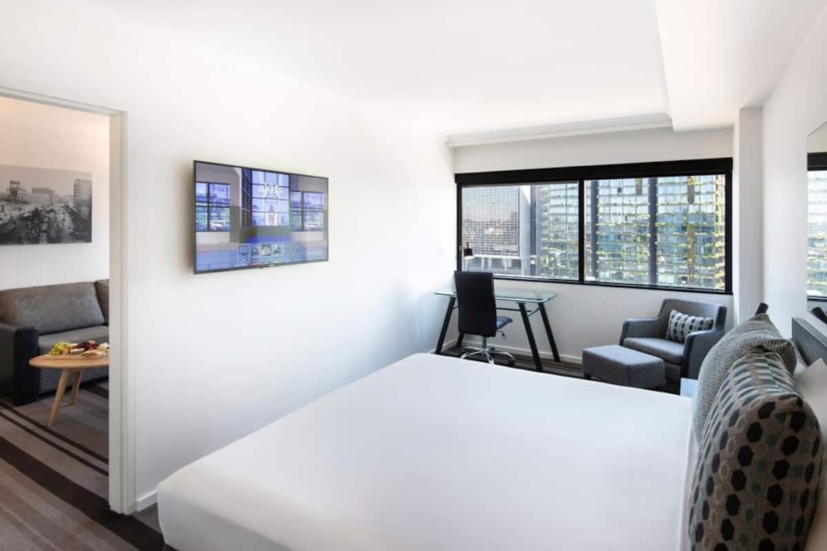 A modern hotel room with a large bed, a desk, a chair, a sofa, and a large window with a city view.