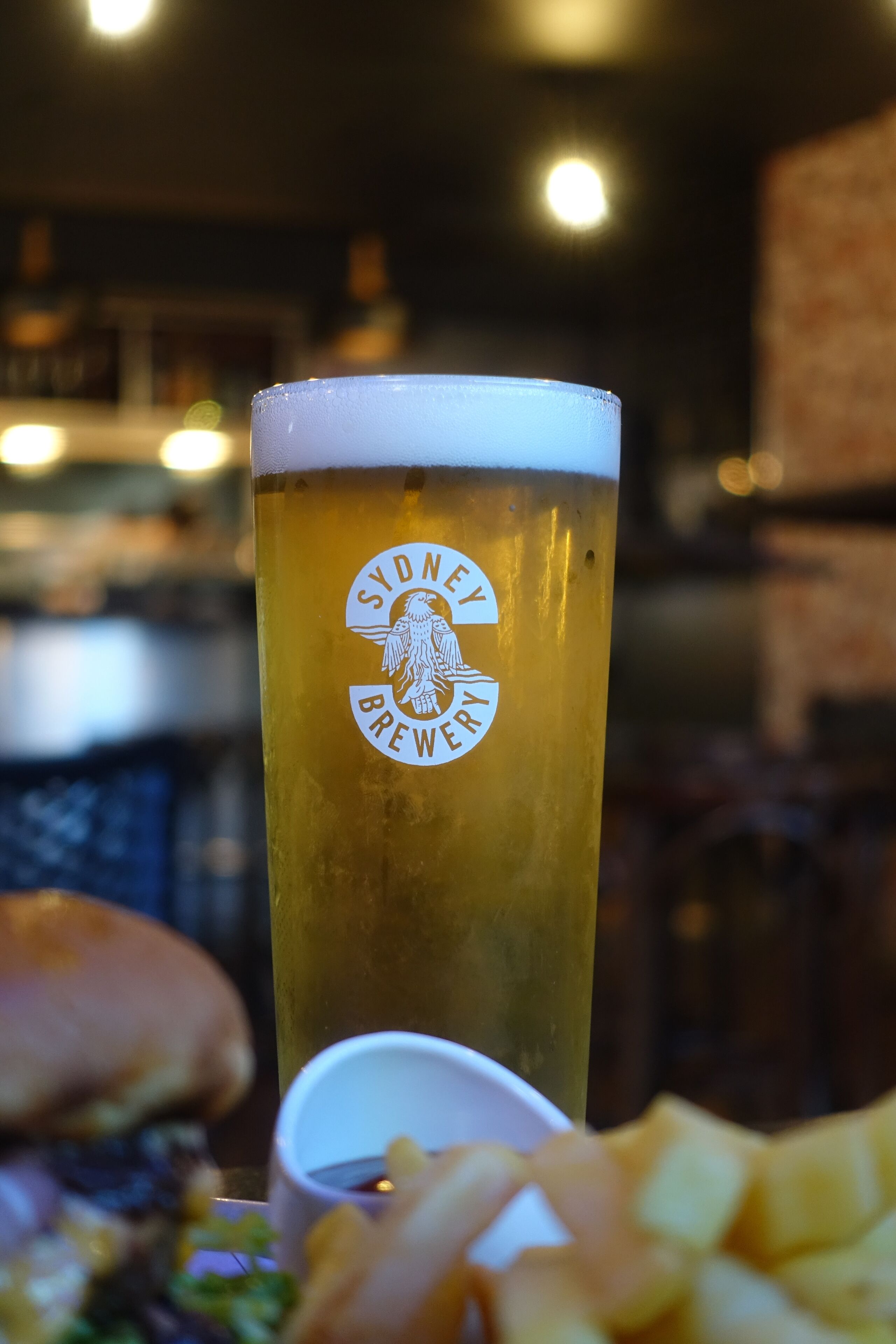 A tall glass of beer with a logo, a burger, and fries on a table.