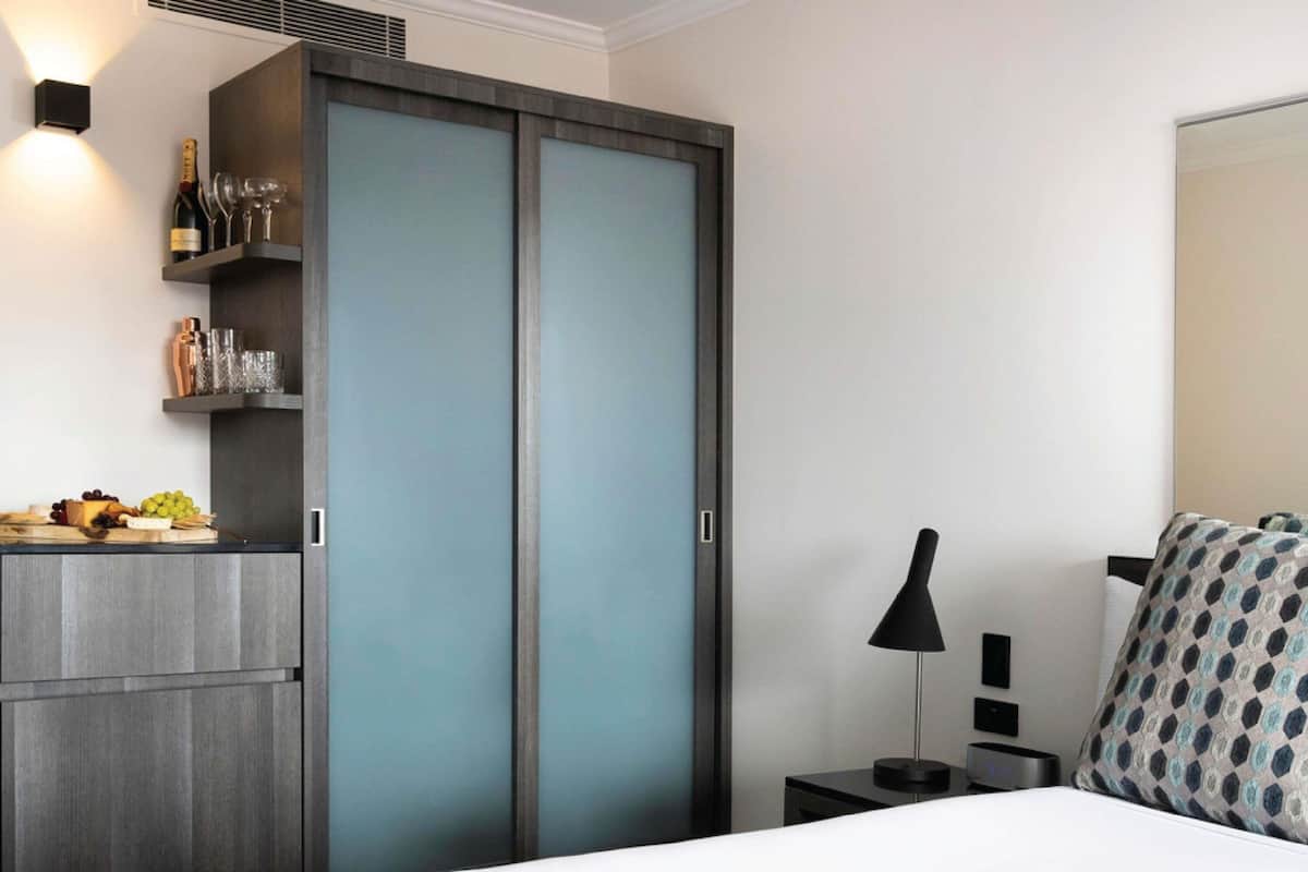 A bedroom with a bed, a nightstand, a lamp, a wardrobe, and a shelf with wine bottles and glasses.
