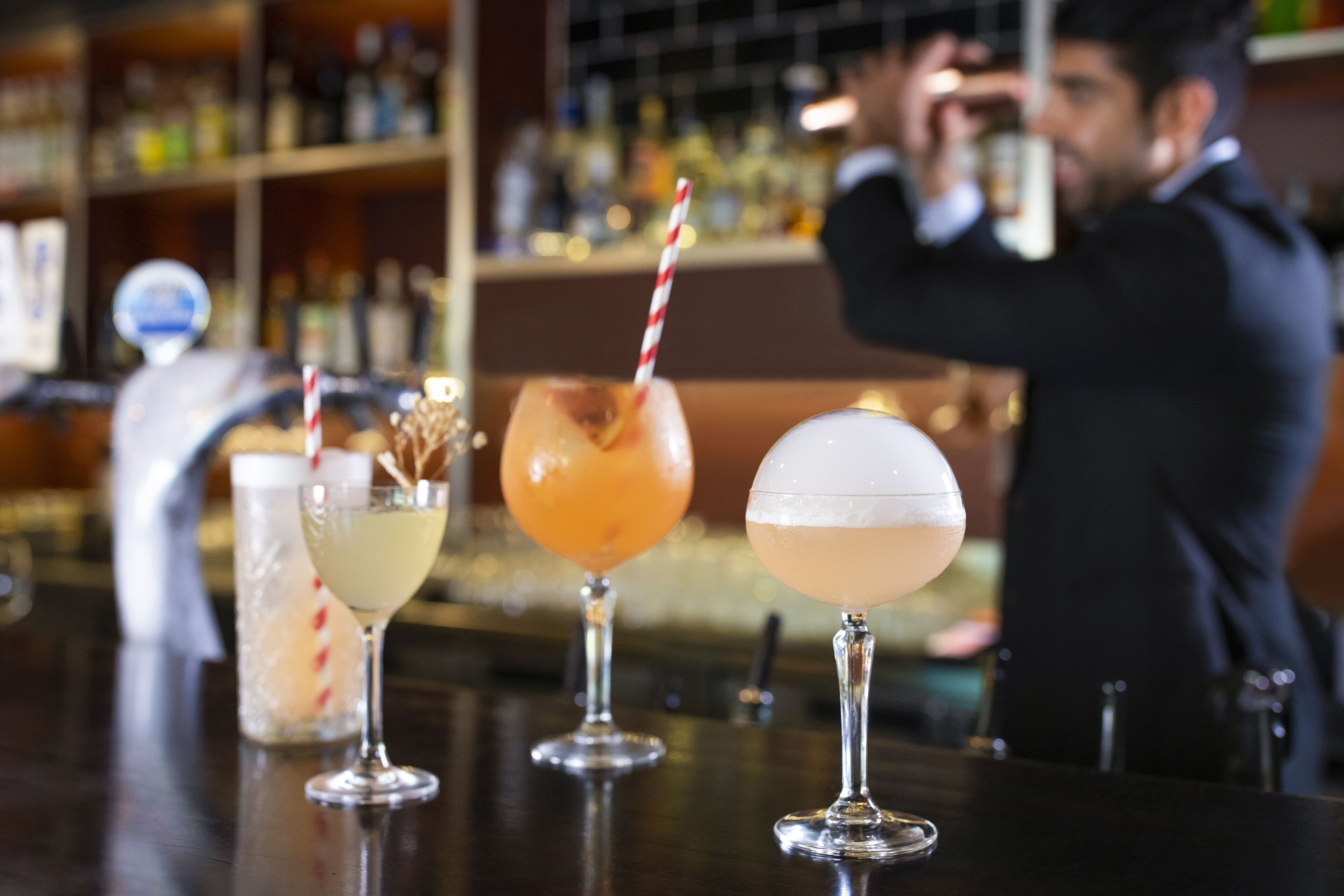 Three cocktails on a bar counter with a blurred background of shelves and a person in the background.