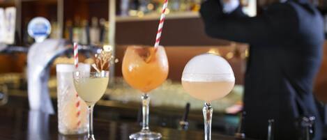 Three cocktails on a bar counter with a blurred background of shelves and a person in the background.