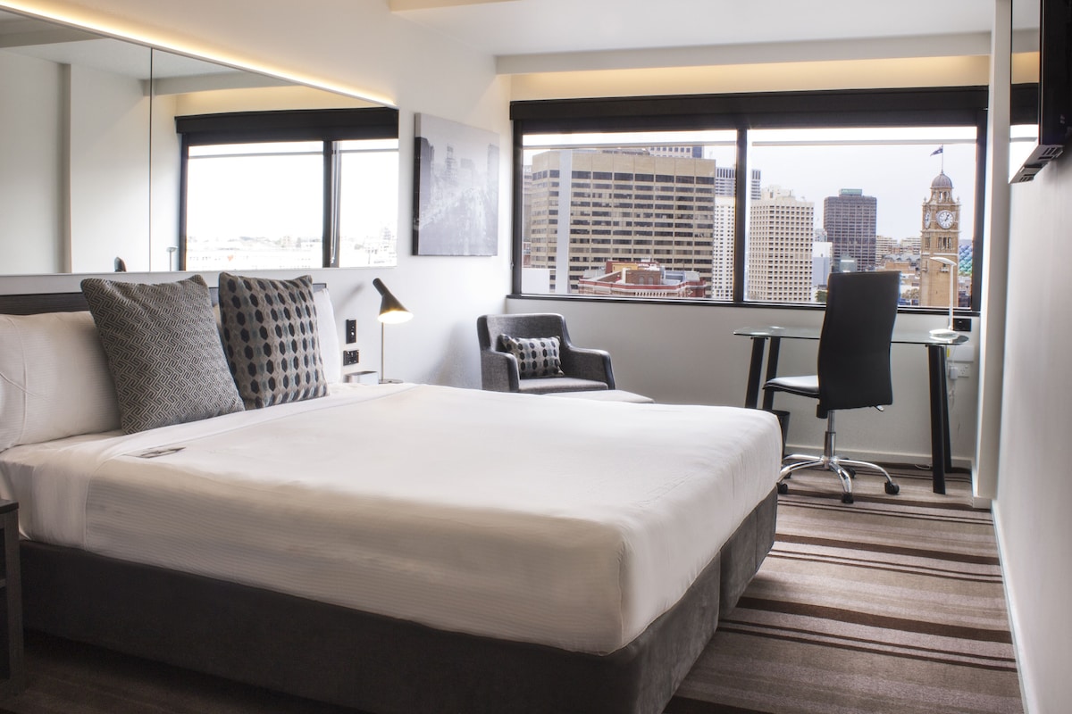 Une chambre d’hôtel avec un grand lit, un bureau, une chaise, une lampe et une vue sur le paysage urbain, avec un beffroi en évidence.