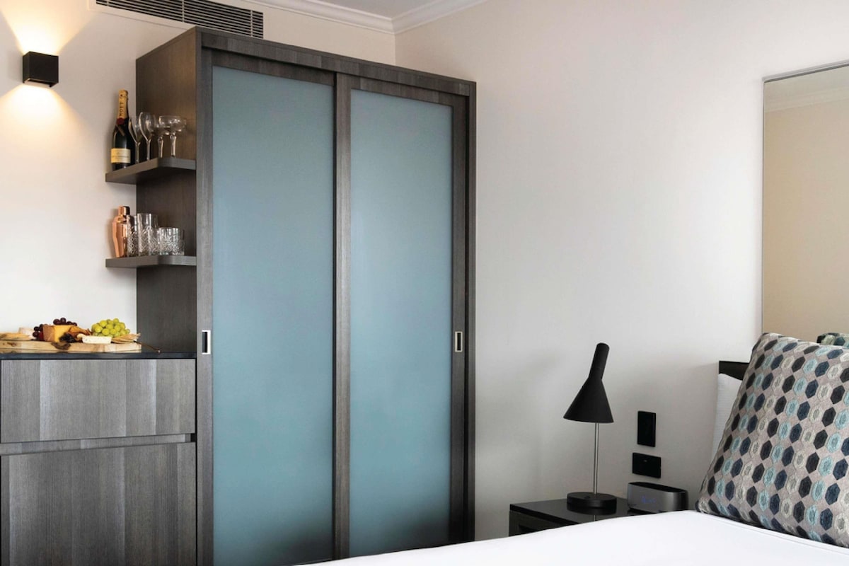 Une chambre à coucher comprenant un lit, une table de chevet, une lampe, une armoire et une étagère avec des bouteilles de vin et des verres.
