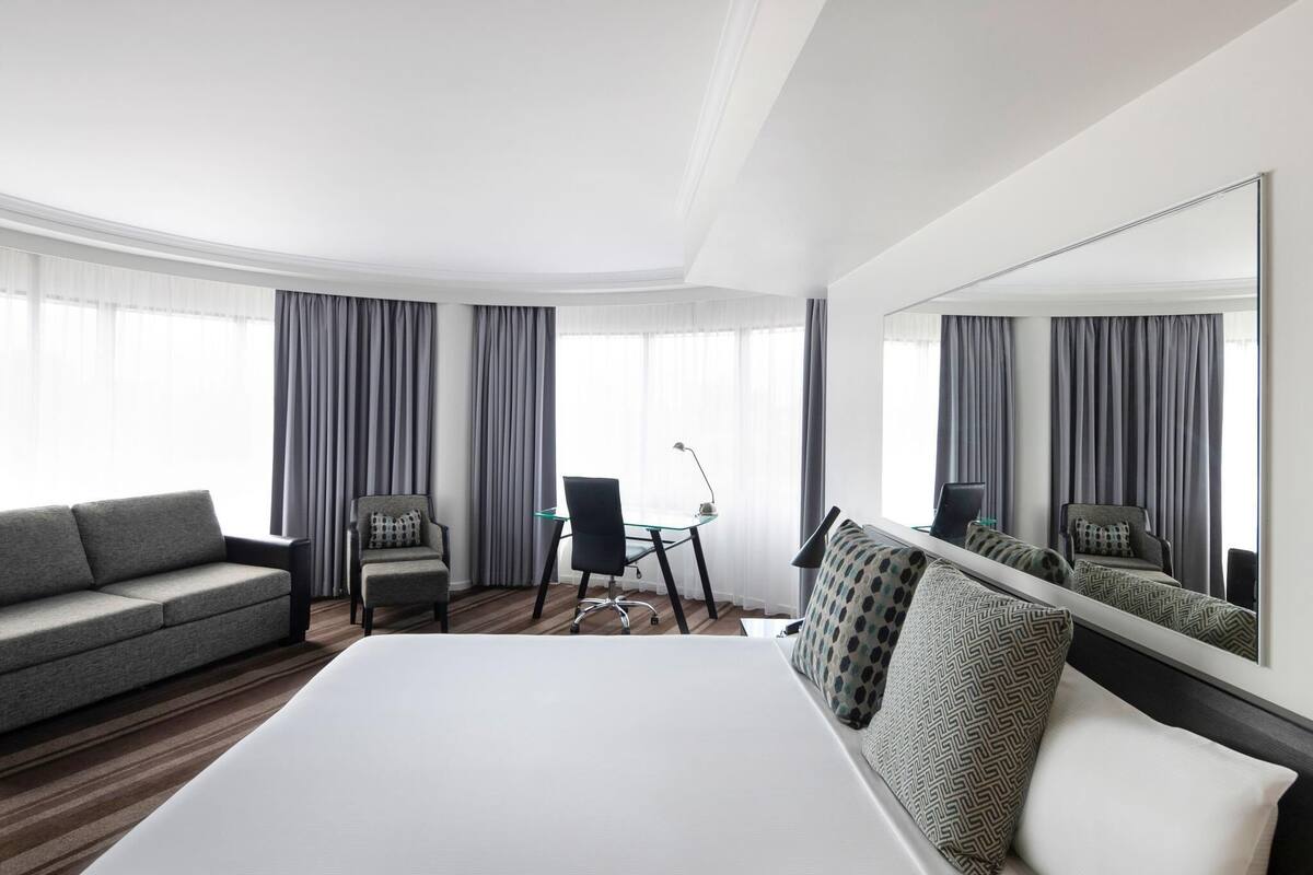 A hotel room with a large bed, a sofa, two armchairs, a desk, and a large mirror.